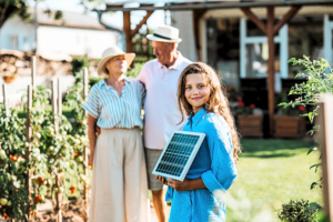 Glückliches Kind mit kleinem PV-Modul in der Hand, steht im Garten vor seinen Großeltern.